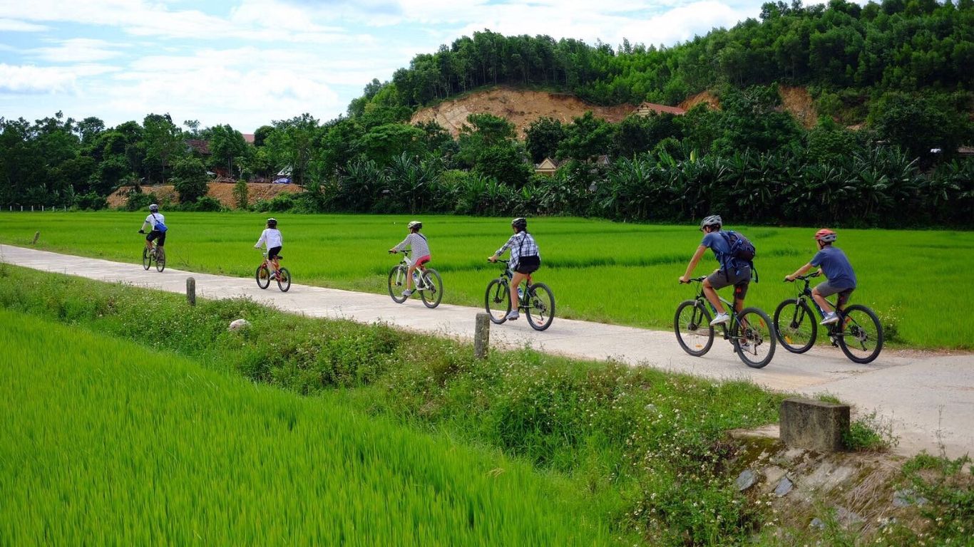 Group of tourists cycling along a narrow rural road through bright green rice paddies in Pu Luong valley, with forested hills and village houses in the background, Thanh Hoa Province, Northern Vietnam.
