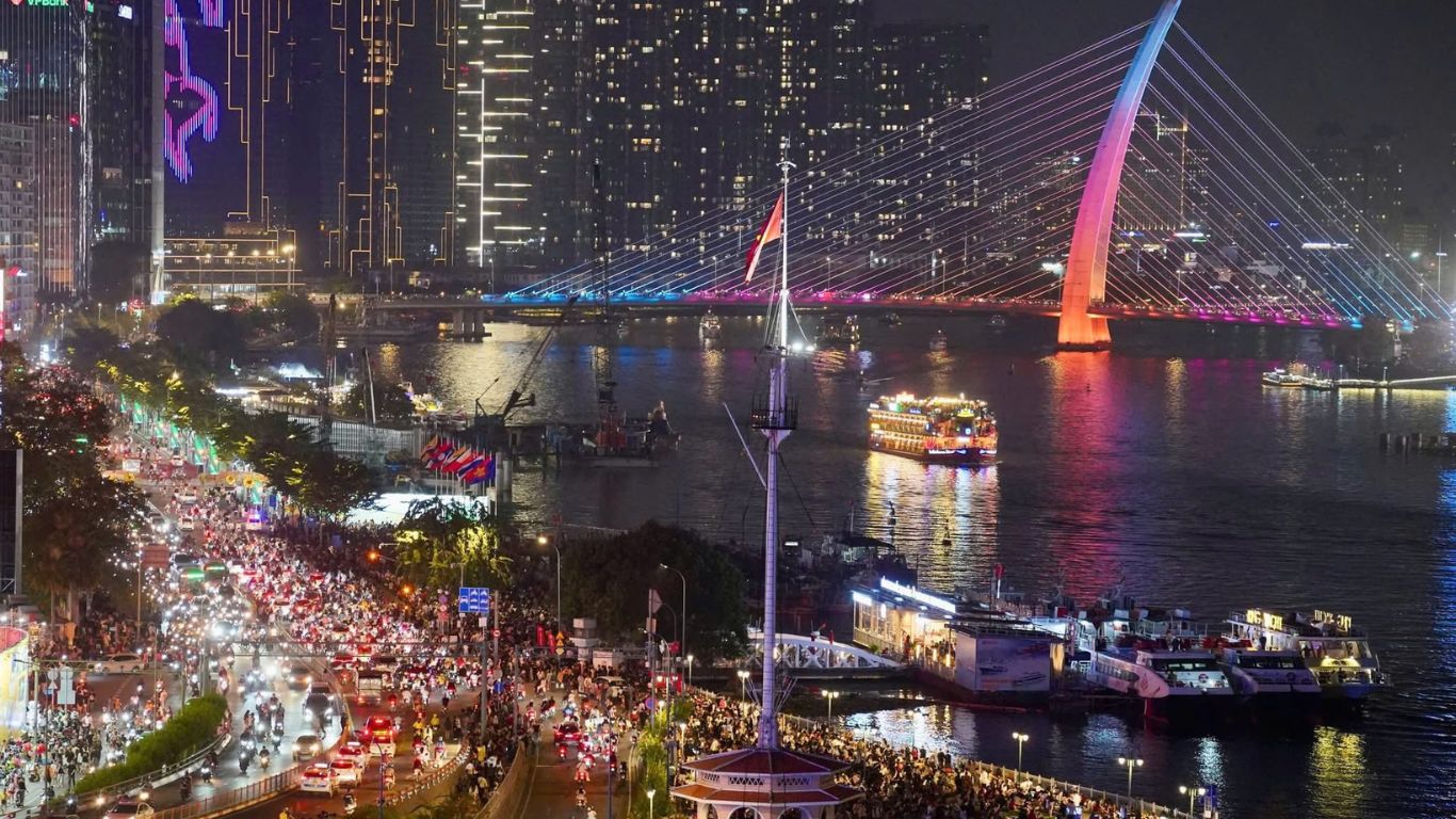 Aerial night view of Nguyen Hue Walking Street in Saigon with dense crowds along the riverside promenade, lit up cruise boats on the Saigon River, and a glowing cable stayed bridge in the background.