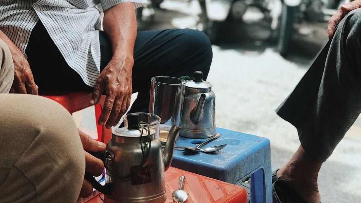 Locals sitting together at a street corner coffee spot in Ho Chi Minh City with traditional metal kettles and glasses on a small plastic stool.