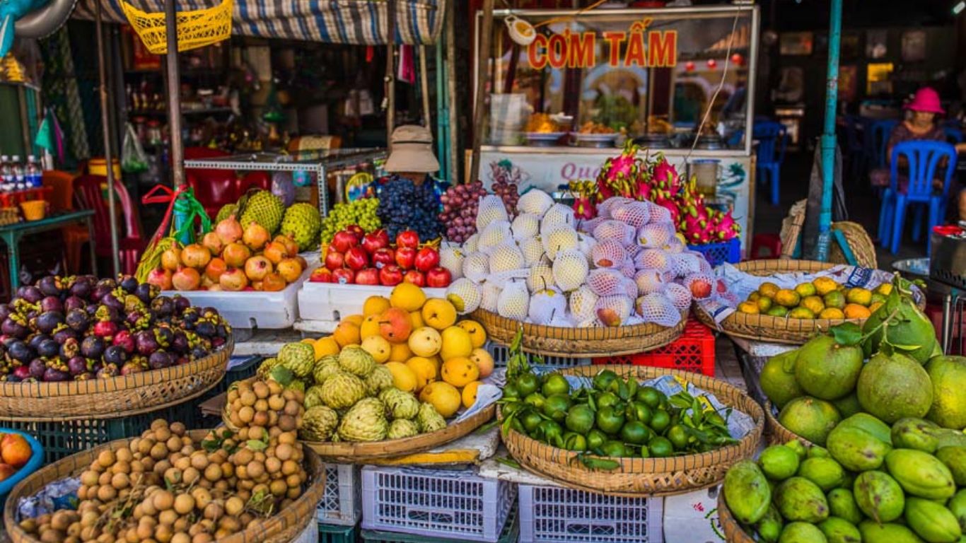 A vibrant street fruit stall in Ho Chi Minh City displaying a colourful spread of tropical fruits including mangosteen, longan, custard apple, dragonfruit, pomelo, and grapes in woven baskets.