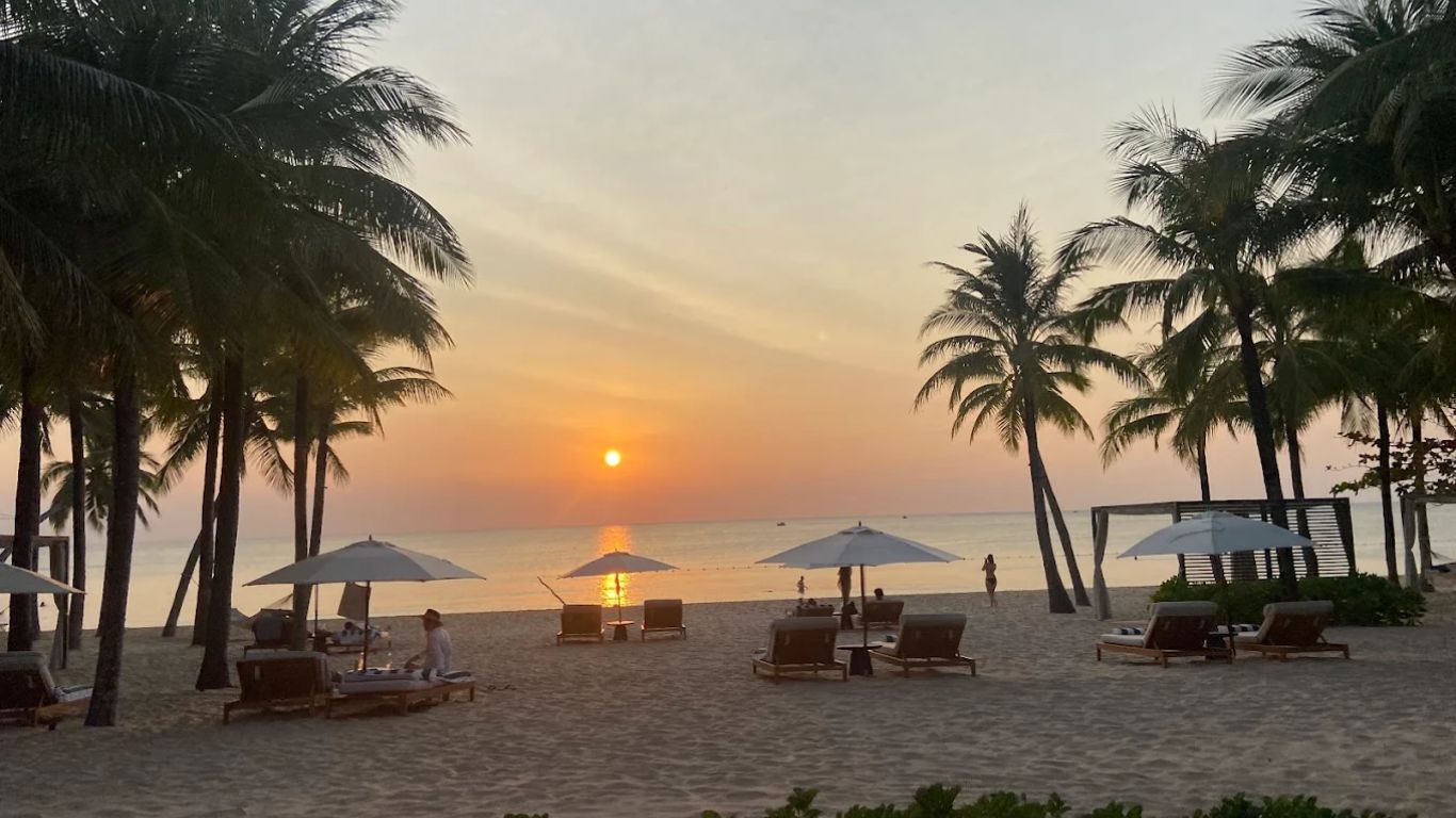 Golden sunset over the sea at a luxury beach resort in Phu Quoc with palm tree silhouettes, white sunbeds and umbrellas on white sand and a glowing orange horizon reflected on calm water