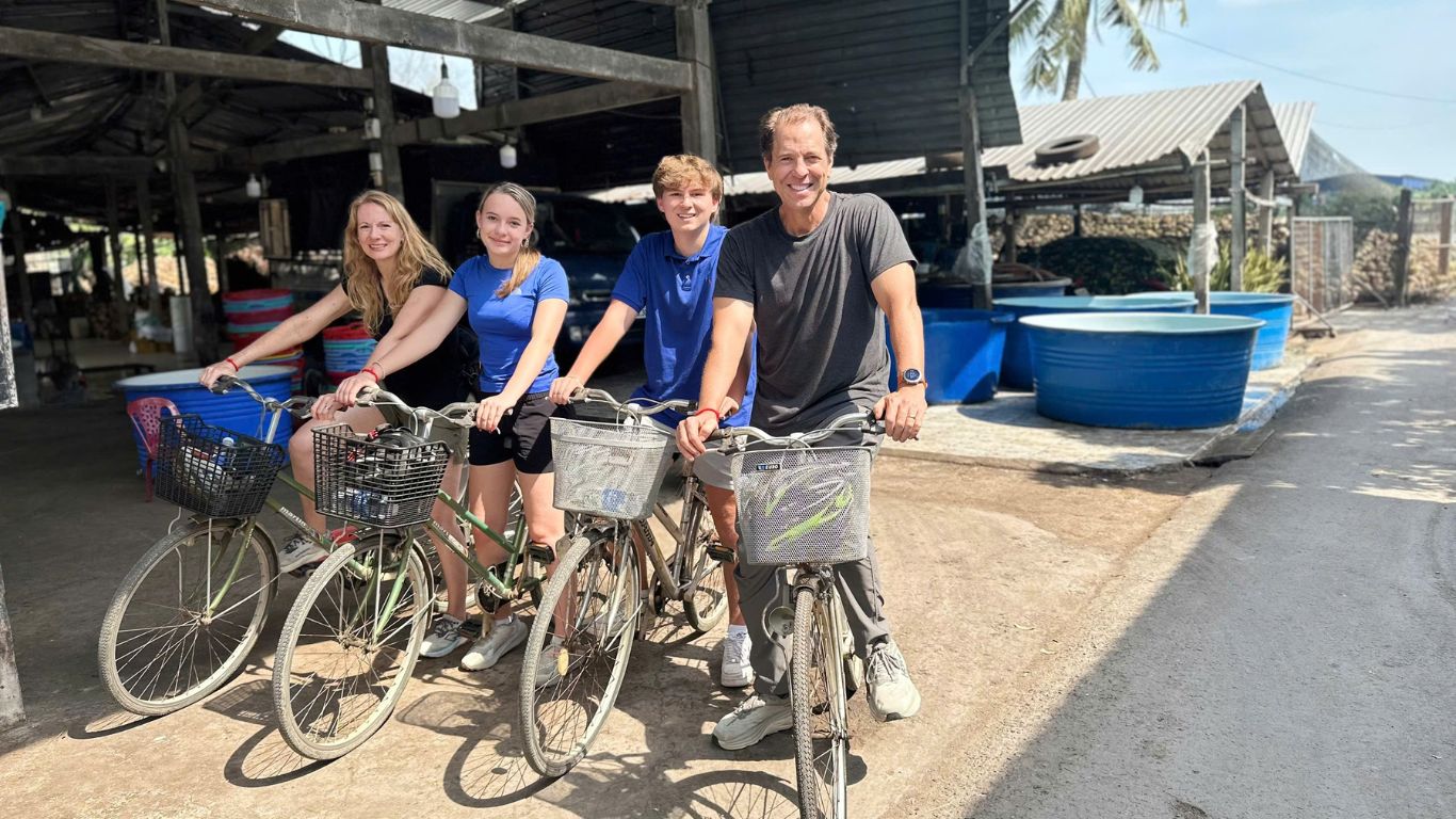 Family of four tourists on bicycles ready to explore a local village in the Mekong Delta, Vietnam, with a traditional workshop and fish farming tanks visible in the background.