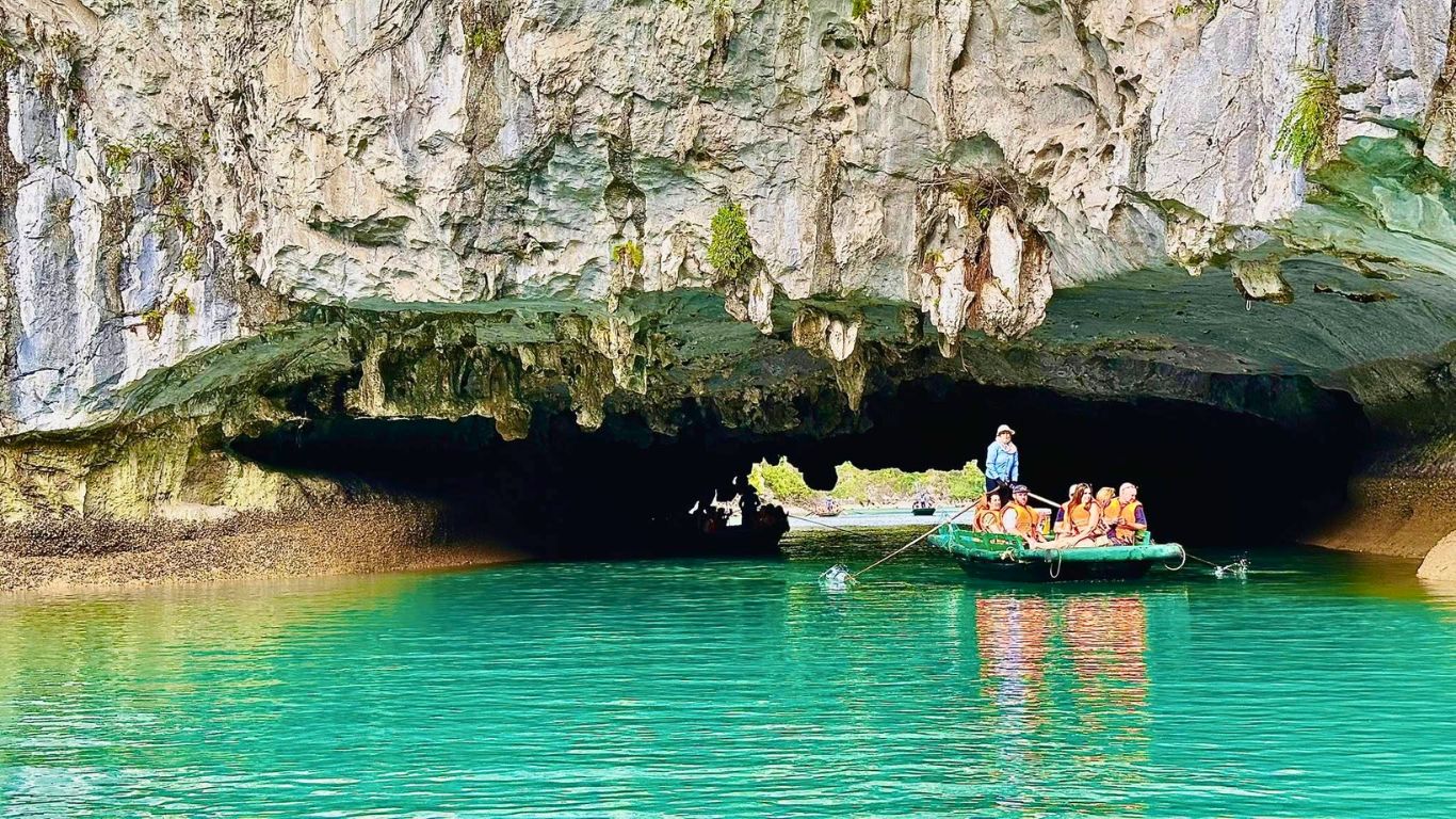 Tourists on a traditional rowing boat entering Dark and Bright Cave in Halong Bay, surrounded by turquoise green water and dramatic limestone cliffs.