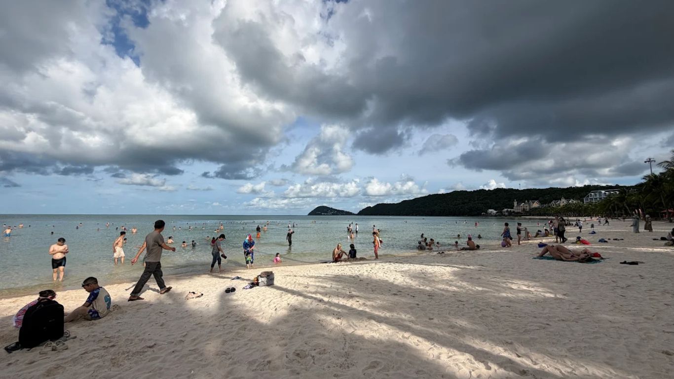 Busy beach in Phu Quoc with swimmers and sunbathers on white sand under dramatic cloudy green season sky with forested hills and resort buildings visible in the background