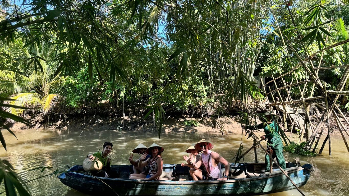 Tourists wearing Vietnamese conical hats on a wooden sampan boat tour through a narrow jungle canal surrounded by bamboo and palm trees in the Mekong Delta, southern Vietnam.