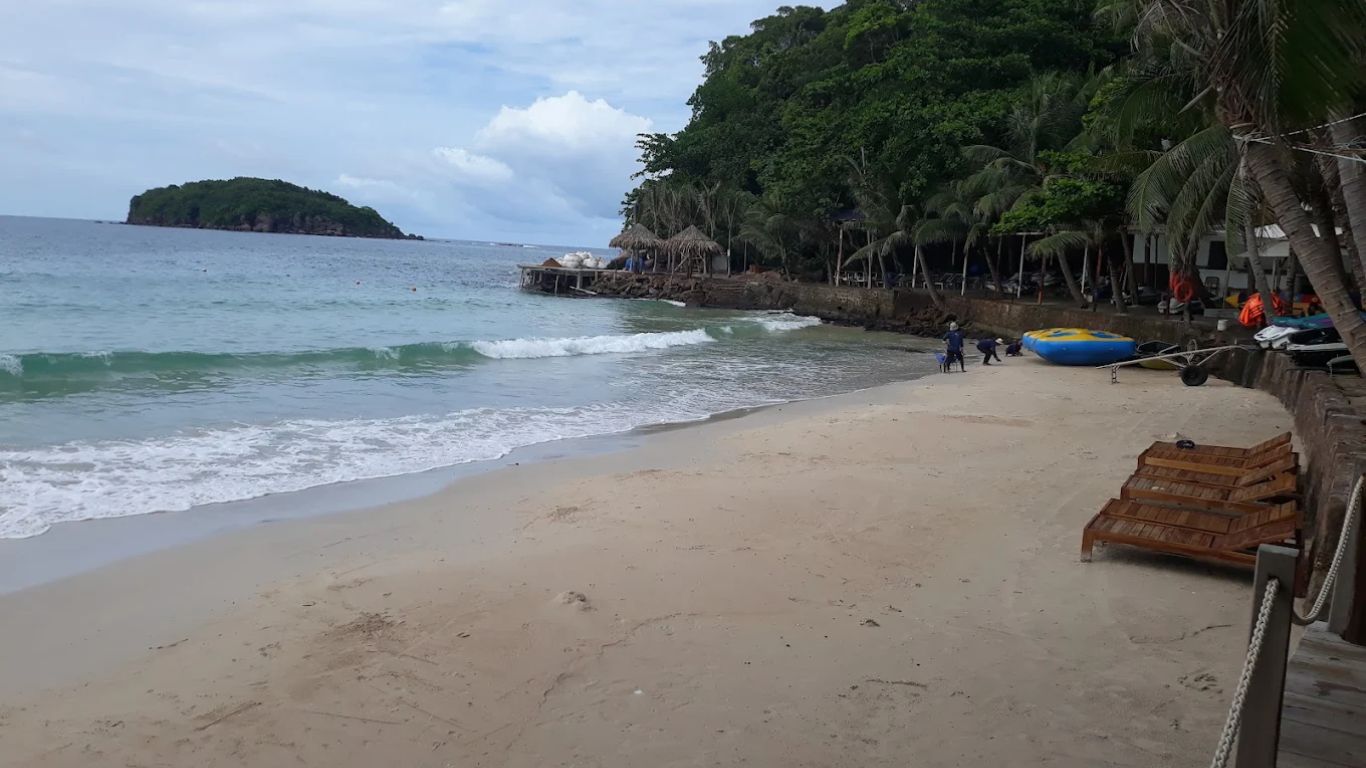 Quiet beach in Phu Quoc during green season with wooden sunbeds, lush tropical trees, turquoise waves, and a small island visible in the overcast distance