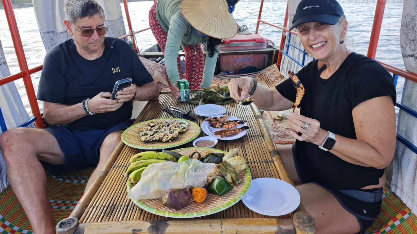 Tourists enjoying a fresh riverside meal on a traditional boat in the Mekong Delta, Vietnam, with grilled prawns, oysters, banana leaf wrapped sticky rice, and local snacks served on bamboo trays by a local host in a conical hat.
