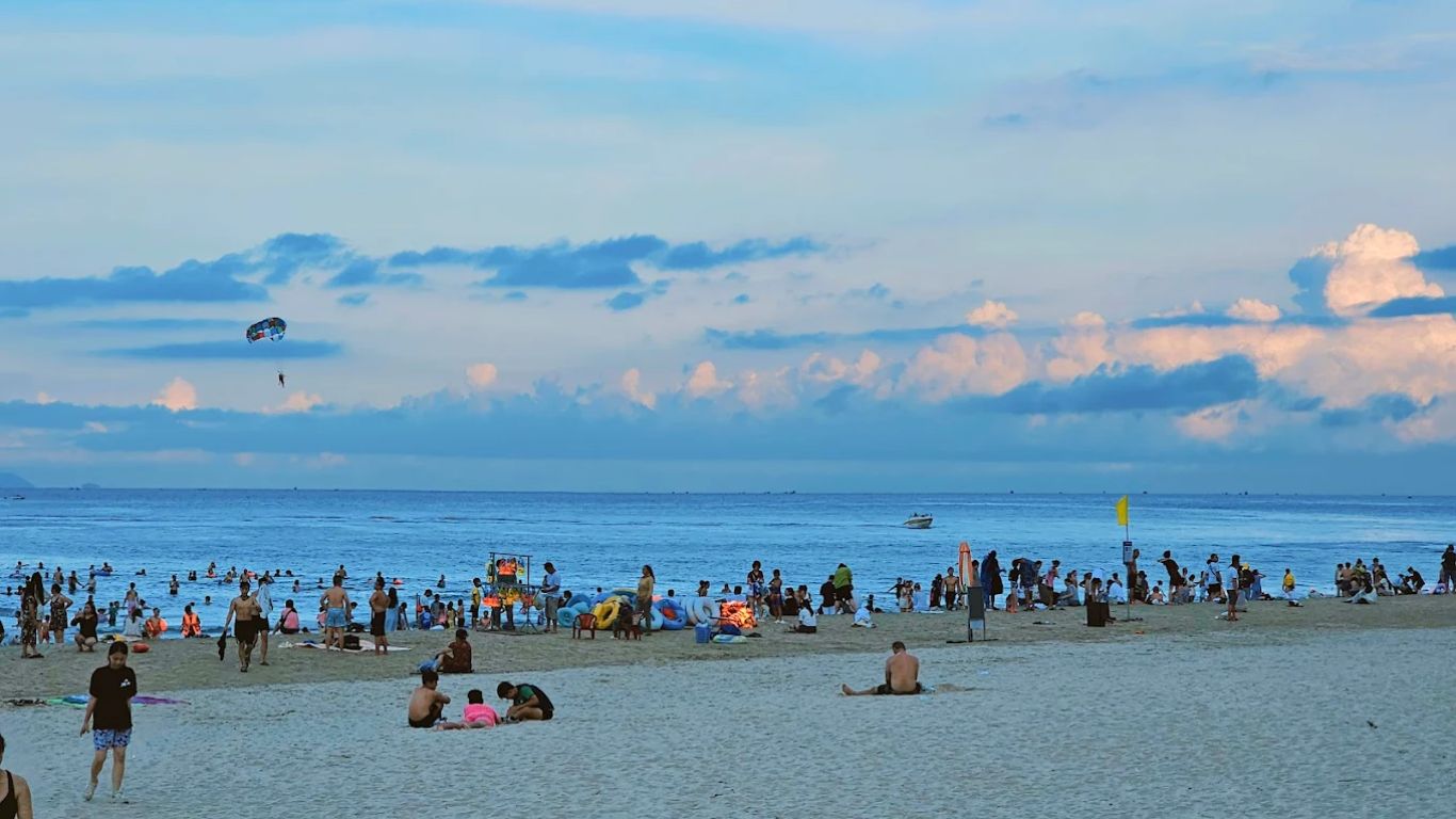 Crowded beach in Da Nang during summer evening with people swimming and parasailing